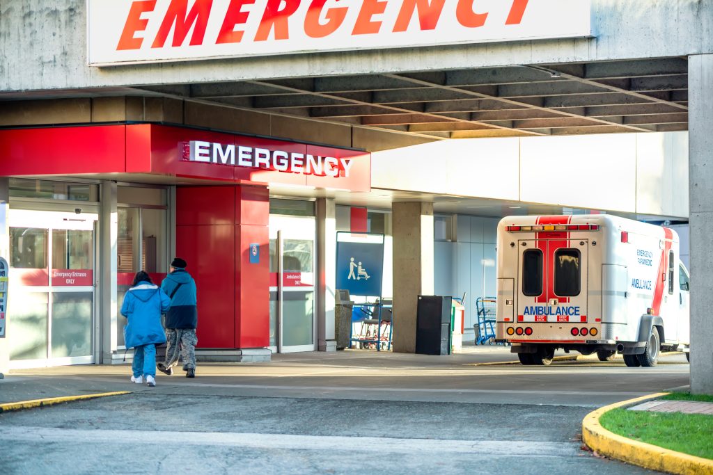 Patients walk into a hospital emergency room entrance while an ambulance stands ready nearby.