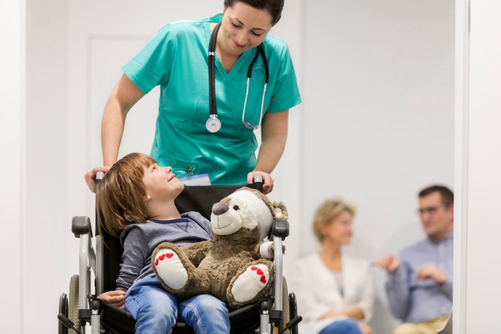Smiling nurse pushing wheelchair with a boy with a teddy bear on it