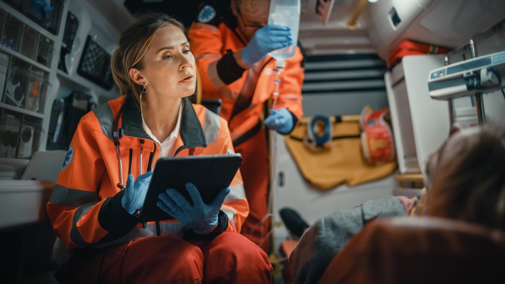 Paramedic in an ambulance listens to a patient while taking notes on a tablet computer.